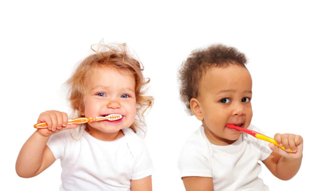 A Caucasian and an African American baby in white undershirts are brushing their teeth with brightly colored toothbrushes.